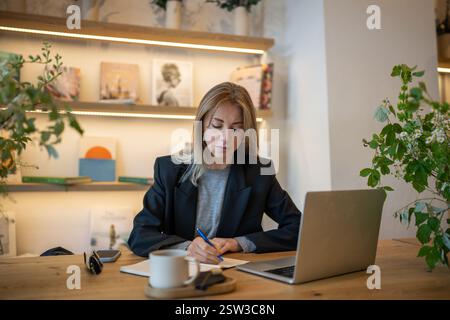 Une femme entrepreneure sérieuse focalisée travaillant avec un ordinateur portable écrit un bloc-notes avec du café dans un lieu de coworking Banque D'Images