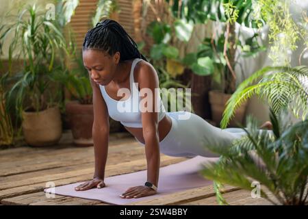 Concentré sur la respiration active femme noire faisant planche sur tapis de fitness avec les yeux fermés dans la chambre avec des plantes Banque D'Images