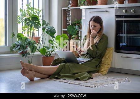Femme inquiète distrait de l'ordinateur portable sur smartphone recevoir de mauvaises nouvelles dans le message assis sur le sol à la maison. Banque D'Images