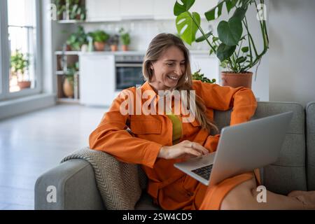 Joyeuse femme d'âge moyen utilisant un ordinateur portable souriant se détendre sur le canapé passer du temps de loisirs à la maison confortable. Banque D'Images