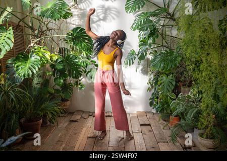 Heureuse femme noire insouciante dansant avec les yeux fermés écoutant de la musique dans des écouteurs parmi les plantes d'intérieur Banque D'Images