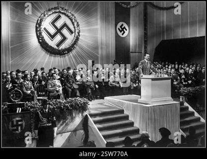 1939 Adolf Hitler sur un podium prononçant un discours lors d'une réunion avec des membres et des fonctionnaires nazis de haut rang. Grand emblème swastika nazi allumé et représenté derrière. Berlin Allemagne nazie.1939 L'année du blitzkrieg allemand en Pologne et le début de la seconde Guerre mondiale Banque D'Images