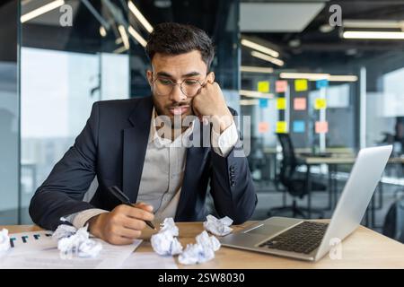 Jeune homme d'affaires fatigué en costume formel assis au bureau avec du papier froissé et un ordinateur portable dans un bureau moderne, se sentant sans inspiration et surchargé de travail Banque D'Images