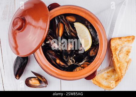 Cuire des moules à la vapeur dans un pot en argile avec du citron et du pain grillé sur une table en bois blanc Banque D'Images