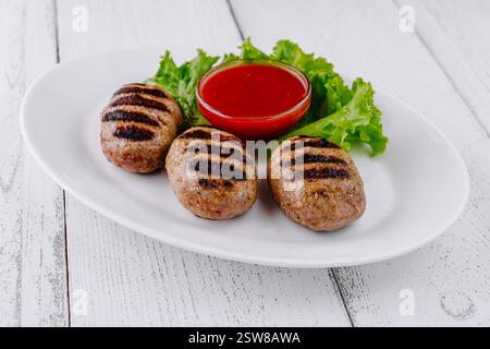 Trois boulettes de viande grillées à la sauce rouge sur une assiette blanche reposant sur une table blanche Banque D'Images