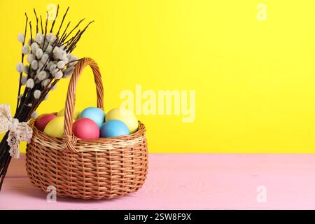 Beaux œufs de Pâques décorés dans un panier en osier et branches de saule sur une table en bois rose sur fond jaune, espace pour le texte Banque D'Images