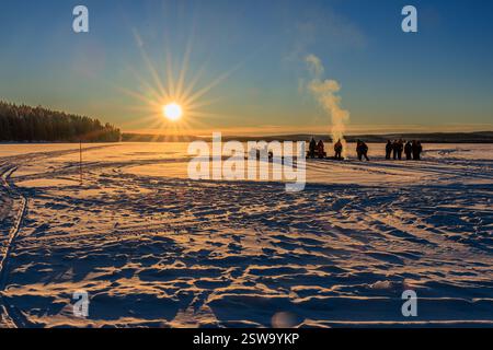 un groupe en silhouette se tient autour d'un feu de camp sur un vaste lac enneigé gelé à côté d'une forêt de pins alors que le soleil se couche bas comme un coup de soleil doré Banque D'Images