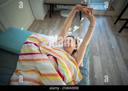 Femme éveillée se trouve dans le lit sous une couverture avec le sourire satisfait s'étirant avec les bras levés. Banque D'Images