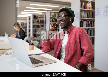Joyeux étudiant afro-américain s'assied dans la bibliothèque à table avec un ordinateur portable et riant Banque D'Images