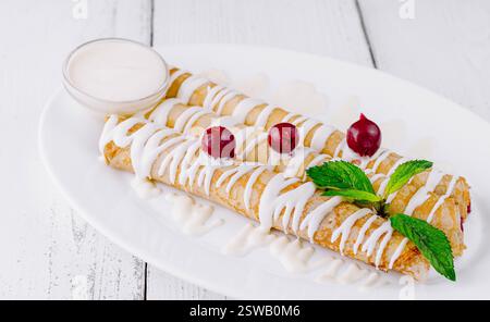 Délicieuses crêpes maison couchées sur une assiette blanche avec de la crème sure et des cerises Banque D'Images