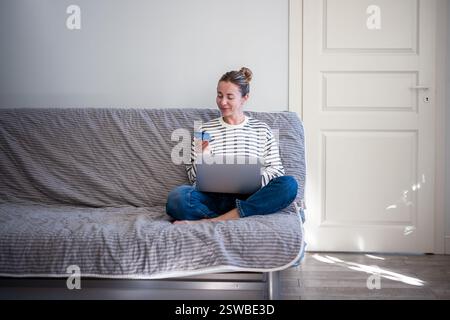 Femme heureuse reposant sur un canapé avec ordinateur portable et carte de crédit. Achats en ligne sur Internet, livraison à domicile. Banque D'Images