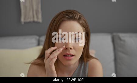 Jeune femme aux cheveux roux à l'intérieur touchant son visage, assise sur un canapé gris dans un salon moderne Banque D'Images