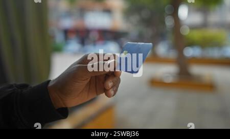 Jeune homme détient une carte de crédit à l'extérieur dans un parc urbain, présentant des méthodes bancaires et de paiement numérique modernes dans un environnement urbain animé avec St floue Banque D'Images