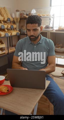 Jeune homme travaillant sur ordinateur portable dans la boulangerie avec des étagères à pain et des pâtisseries en arrière-plan Banque D'Images