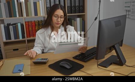 Jeune femme travaillant à un bureau dans une pièce de bureau avec des papiers, un ordinateur et des étagères pleines de livres et de classeurs en arrière-plan Banque D'Images