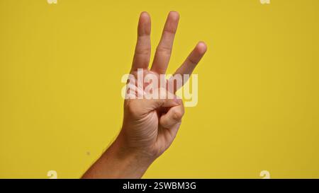 Gros plan de la main d'un homme montrant trois doigts sur un fond jaune vif, symbolisant le chiffre trois ou un compte à rebours Banque D'Images