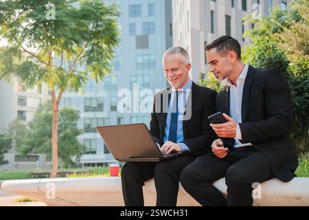 Deux hommes d'affaires confiants se sont engagés dans une discussion productive tout en utilisant un ordinateur portable à l'extérieur. Leur tenue professionnelle reflète leur dévouement au travail d'équipe et à la collaboration, leurs rôles en tant que cadres. Photo de haute qualité Banque D'Images