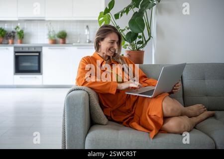 Joyeuse femme d'âge moyen utilisant un ordinateur portable souriant se détendre sur le canapé passer du temps de loisirs à la maison confortable. Banque D'Images
