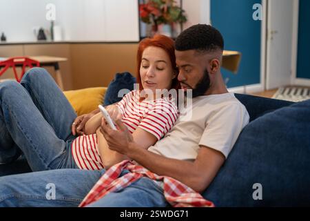 Jeune millénaire couple divers homme et femme se reposant à la maison avec le téléphone mobile Banque D'Images