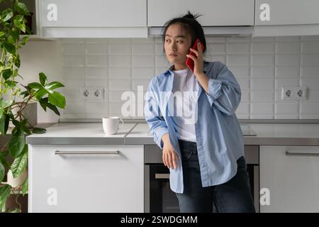 Femme asiatique pensive fatiguée debout dans la maison à la cuisine avec une tasse de café faisant appel dans le téléphone portable Banque D'Images