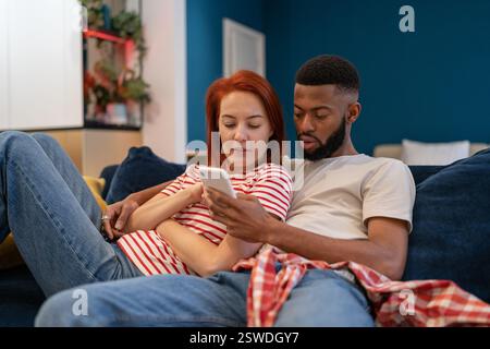 Jeune millénaire couple divers homme et femme se reposant à la maison avec le téléphone mobile Banque D'Images