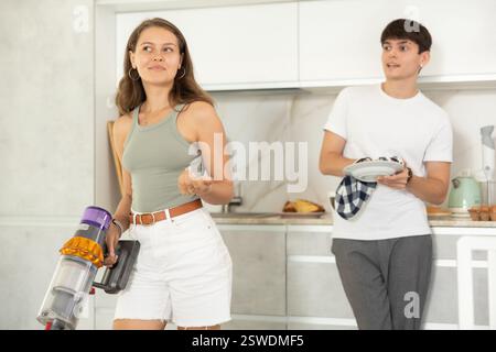 Jeune femme aspirant dans la cuisine tout en communiquant avec son petit ami séchant la vaisselle avec une serviette Banque D'Images
