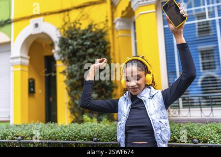 Heureuse femme afro-américaine dansant levant les bras tout en écoutant de la musique avec des écouteurs et un téléphone portable dans la ville dans une rue colorée Banque D'Images