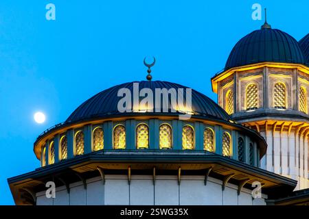 Une vue rapprochée des dômes d'une mosquée au crépuscule, avec des détails architecturaux complexes et des fenêtres éclairées. La lune est visible en clair Banque D'Images
