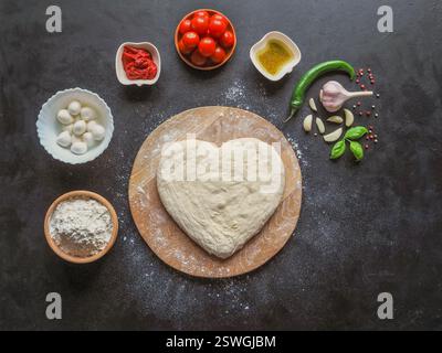 Pâte en forme de coeur et un ensemble d'ingrédients pour pizza sur une table noire. La vue depuis le haut Banque D'Images