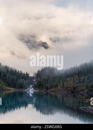 Paysage alpin atmosphérique avec des arbres près du lac de montagne et haute montagne enneigée dans les nuages bas. Vue verticale. Banque D'Images