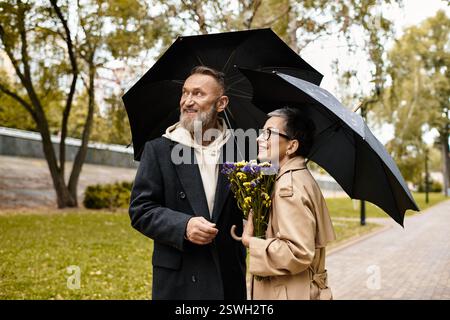 Un couple se promène dans un parc verdoyant, partageant la joie sous des parapluies avec des fleurs à la main. Banque D'Images