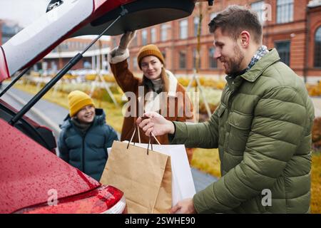 Famille avec enfant emballant le coffre de voiture avec des sacs à provisions dans un cadre urbain extérieur pendant l'automne, parents souriants et enfant vêtus de vêtements chauds et de chapeaux profitant du temps en famille Banque D'Images