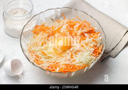 Un bol en verre rempli de chou blanc râpé et de carottes râpées, avec un œuf cru pour une recette de crêpes aux légumes maison ou de fritter entourée Banque D'Images