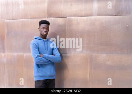 Un jeune homme dans un sweat à capuche bleu se tient devant un mur. Il regarde la caméra les bras croisés. La scène est sérieuse et contemplative Banque D'Images