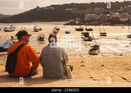 Vue sur Harbour Beach, St Ives, Cornwall, Angleterre à marée basse avec, de petits bateaux et un homme et une femme assis sur le quai relaxant Banque D'Images