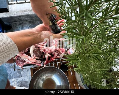 Une personne assaisonne les côtelettes d'agneau avec du poivre noir tout en se préparant pour un barbecue à l'extérieur. Le romarin frais ajoute de la saveur. Banque D'Images