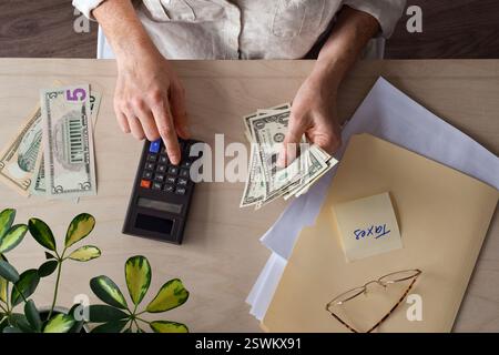 Vue de haut en bas d'une femme assise à un bureau comptant des dollars américains en argent comptant avec une calculatrice faisant des impôts Banque D'Images