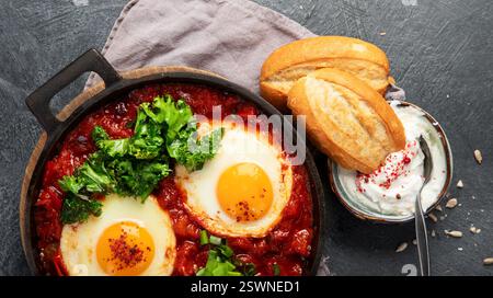 Oeufs Shakshuka dans une casserole avec du pain sur un fond sombre. Oeufs pochés dans une sauce épicée au poivre tomate. Œufs brouillés juifs traditionnels. Vue de dessus. PA Banque D'Images