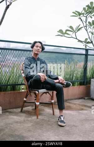 Jeune homme élégant se relaxant sur une chaise sur le toit. Il porte une tenue décontractée et dégage une ambiance contemplative. L'arrière-plan du paysage urbain ajoute un sen Banque D'Images