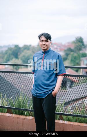 Portrait d'un jeune homme asiatique souriant en tenue décontractée, debout à l'extérieur avec un décor pittoresque. Il respire la confiance et un comportement détendu, showca Banque D'Images