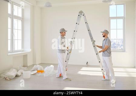 Peintres de maison debout dans une pièce vide près de l'échelle se préparant à peindre le mur du nouvel appartement. Banque D'Images