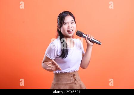 Une jeune femme, avec un micro, chantant avec passion, sourit et apprécie sa performance. isolé sur fond orange Banque D'Images