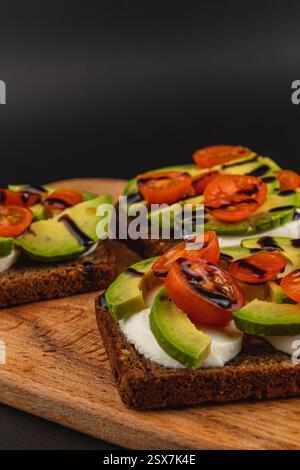 Les toasts sont magnifiquement disposés sur une planche de bois, surmontés de fromage crémeux, de tranches d'avocat mûres et de tomates cerises colorées. Un filet de balsamique Banque D'Images