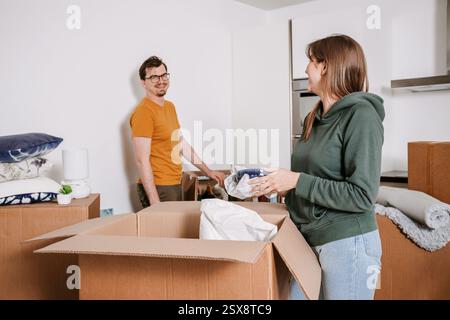 Une femme déballe les ustensiles de cuisine d'une boîte tandis que son partenaire sourit en arrière-plan. Leur nouvelle maison est remplie de boîtes mobiles Banque D'Images