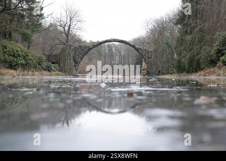 Kromlau, Allemagne. 23 février 2025. Le pont de Rakotzbrücke se reflète dans un lac couvert de glace dans le parc Azalée et Rhododendron. Crédit : Sebastian Kahnert/dpa/Alamy Live News Banque D'Images