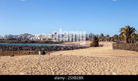 Vue sur la plage de sable ensoleillée avec des gens se relaxant et une ville côtière en arrière-plan sous un ciel bleu clair Costa Teguise Lanzarote Banque D'Images