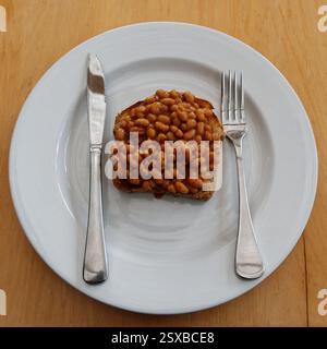 Haricots cuits sur du pain grillé prêt à être mangé. Ils sont sur une assiette blanche avec un couteau et une fourchette. Ils ont une aspersion de poivre noir sur le dessus. Banque D'Images