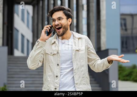 Appel téléphonique, bonnes nouvelles, potins. Heureux homme indien surpris dans une conversation agréable sur smartphone, profiter de parler avec un ami à l'extérieur. Un homme hindou arabe debout dans la rue de la ville. Les modes de vie de la ville Banque D'Images