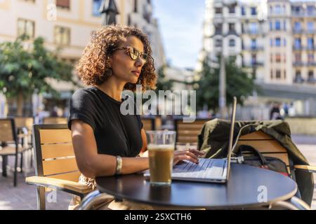 Vue latérale d'une femme d'affaires arabe confiante en beauté travaillant avec un ordinateur portable et buvant du café à l'extérieur Banque D'Images
