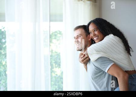 Couple interracial heureux plaisantant regardant par une fenêtre à la maison ou chambre d'hôtel Banque D'Images